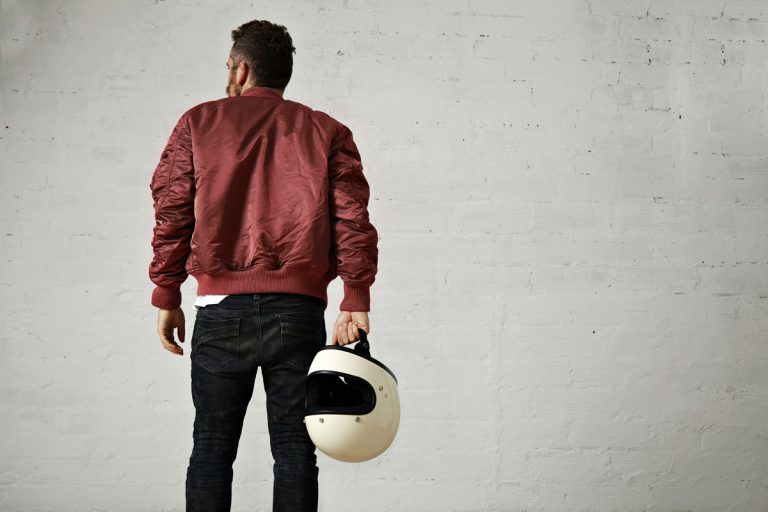 Man wearing a bomber jacket and holding a helmet while staring at a gray wall, Are Bomber Jackets Warm Enough For Winter?