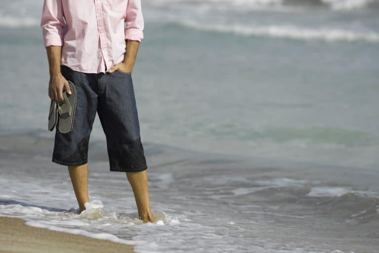 A man wearing a white top and semi wet Capri pants while standing on the shoreline while holding his sandals, Should Guys Wear Capri Pants?