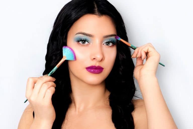 A beautiful woman putting on blue eyeshadow on her eyebrows while holding make up brushes on both her hands, What Color Lipstick Goes With Blue Eyeshadow?