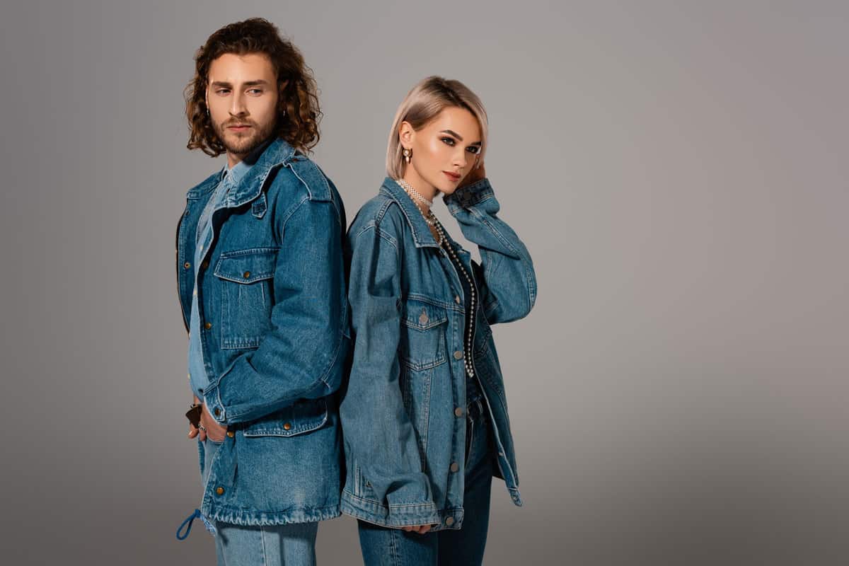 A couple wearing long denim jackets and jeans while standing on a gray background