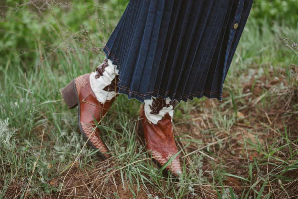 A woman wearing cowboy boots and pleated denim skirt while standing on grass