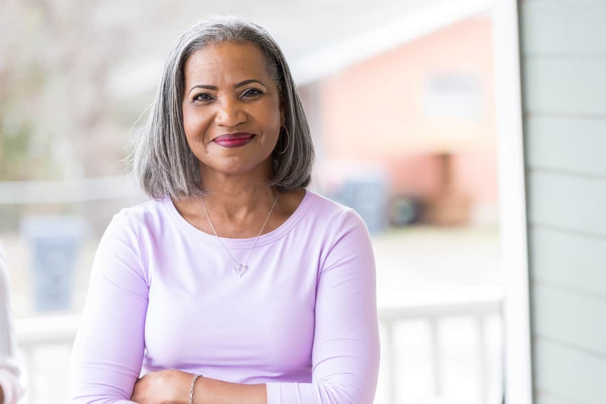 Beautiful African American senior woman with makeup stands confidently on her front porch. She is smiling at the camera.