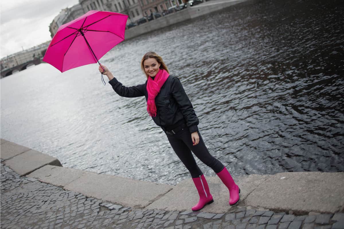 Beautiful young and happy blond woman in a bright pink scarf, rubber boots and umbrella walking in a rainy city