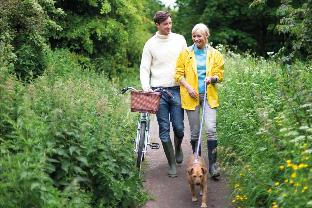 Couple out walking a small brown dog along a dirt path in the countryside wearing rain boots