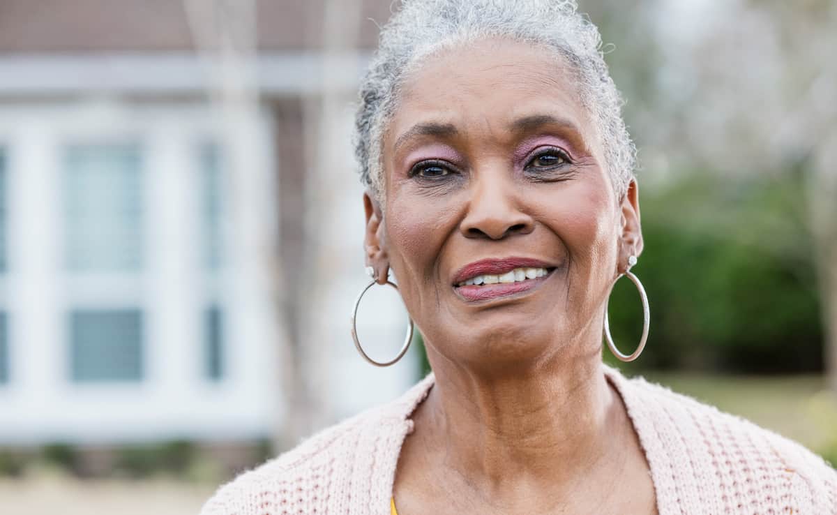 The-face-of-a-senior-African-American-woman-with-short,-gray-hair,-standing-outside-her-home.-She-is-smiling-and-looking-toward-the-camera.
