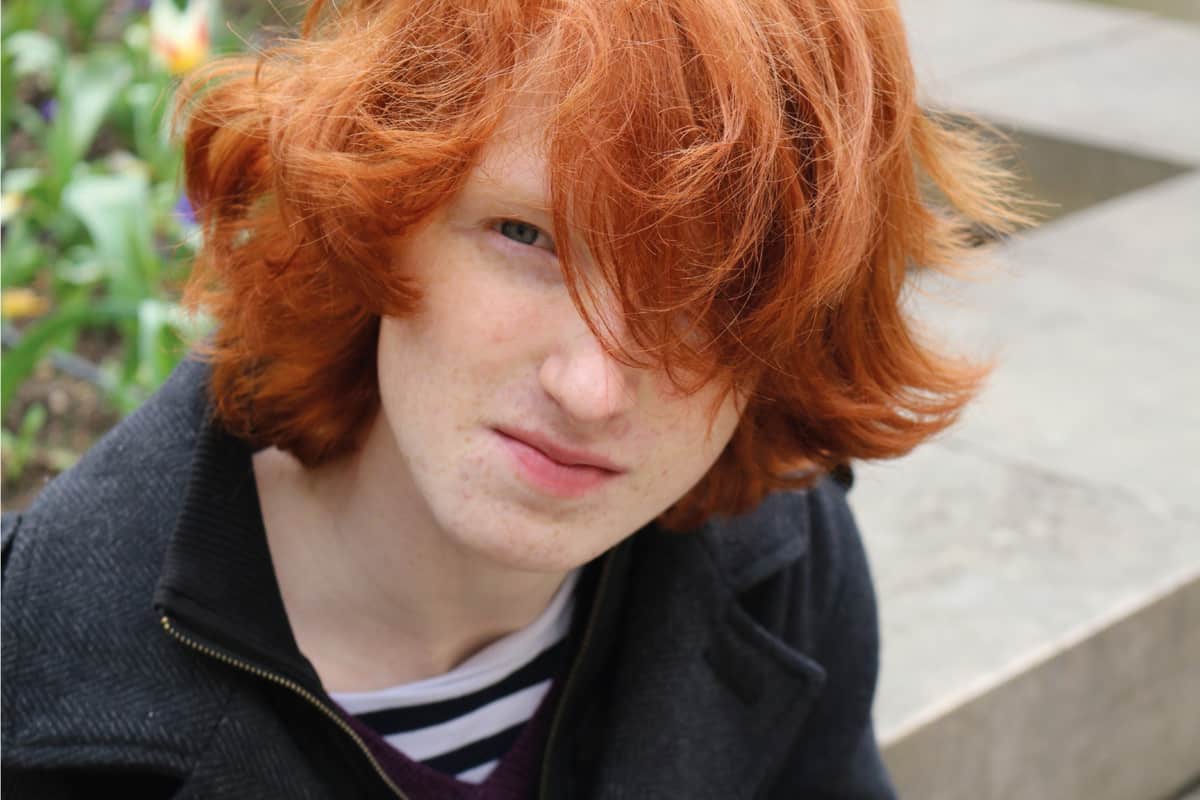 ginger haired teenager squinting through overgrown fringe at the camera whilst sat outside