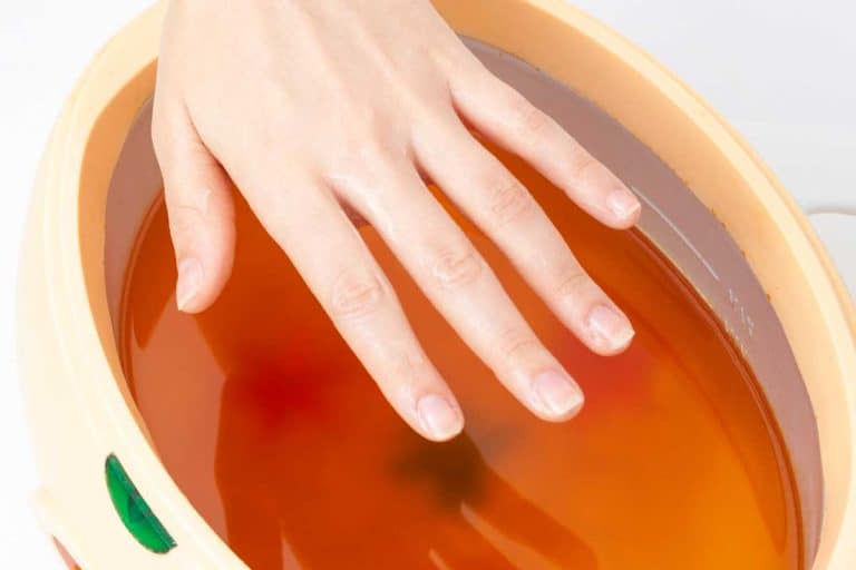 A female hand and orange paraffin wax bowl, How To Do Paraffin Manicure At Home [Even Without A Machine]