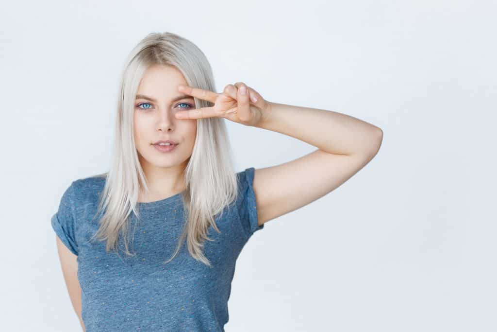portrait of a beautiful girl student with blond hair wearing clean t-shirt showing peace gesture isolated on grey background. Amazing blue eyes and grey dyed hair.
