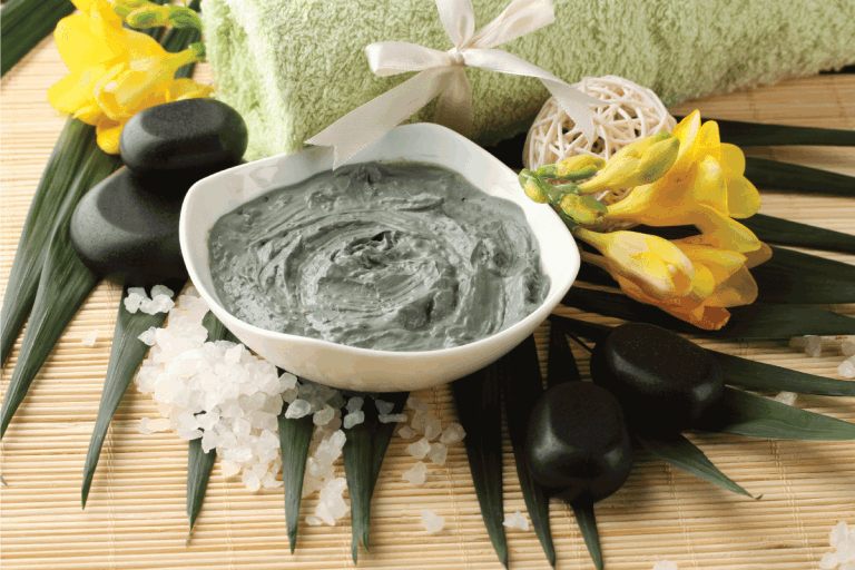 green colored cosmetic clay in a ceramic bowl on a bamboo mat with hot stone and towel. How Long Should You Keep A Clay Face Mask On