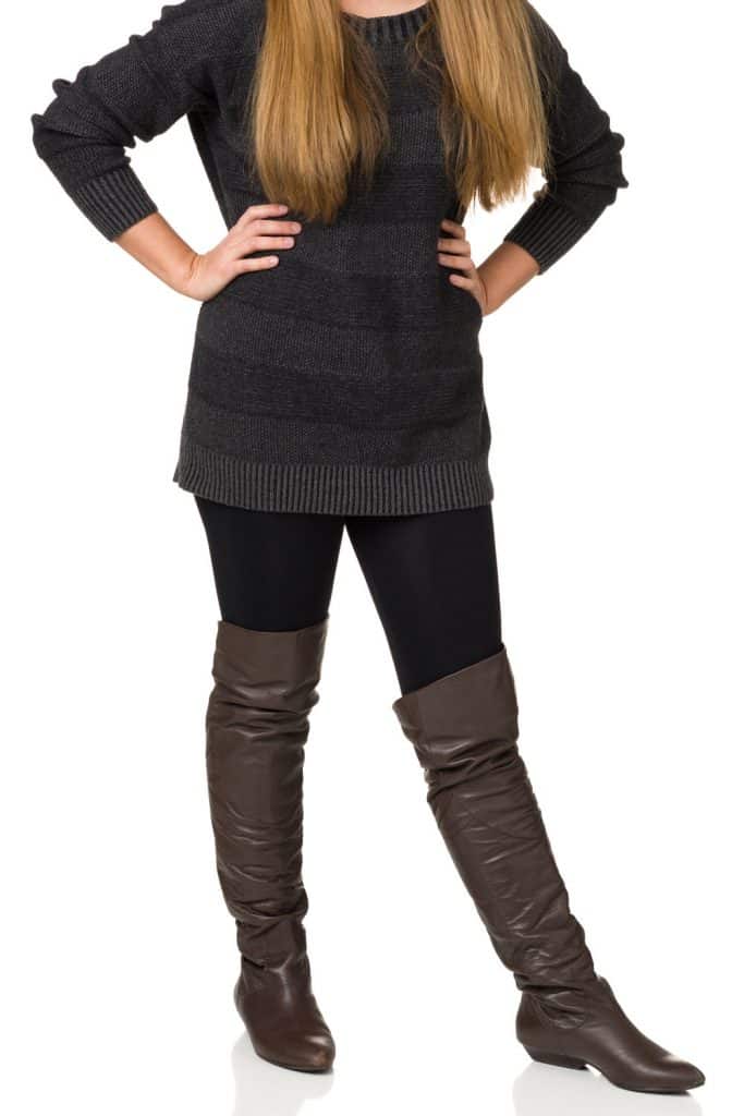 A woman wearing a sweater, black leggings, and brown long boots