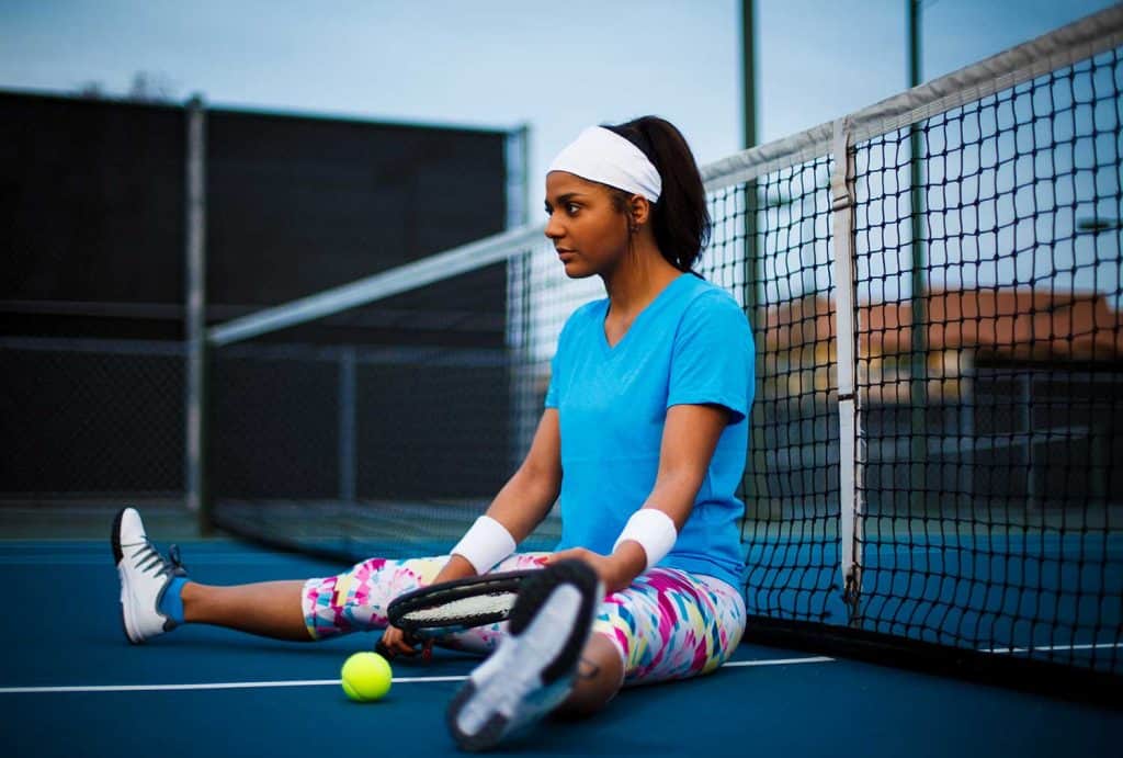A young woman sits and rests at the center of a tennis court next to the net