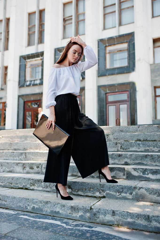Portrait of a flawless young woman in white blouse, wide black pants and classic high heels posing on the stairs