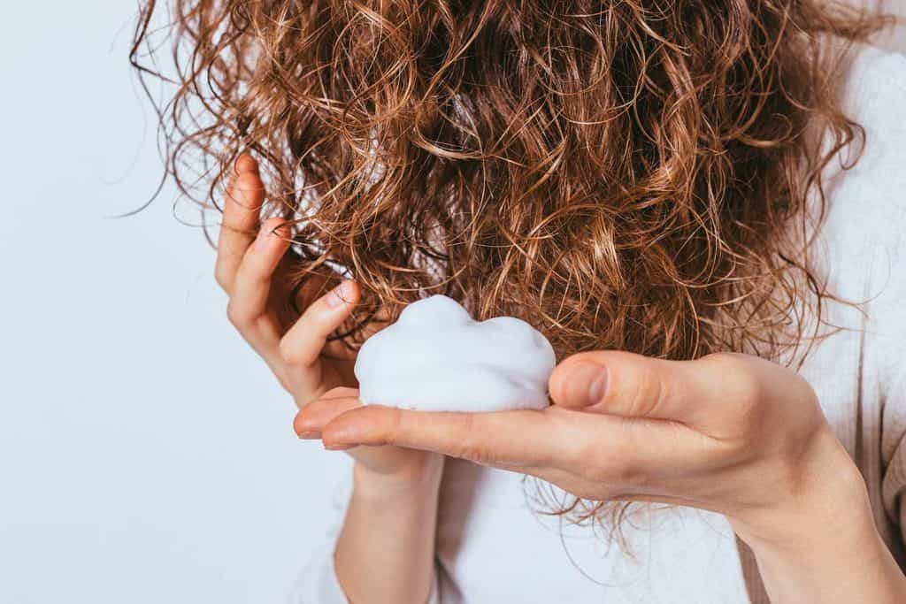 Woman's hands apply styling mousse to her curly hair on white studio background