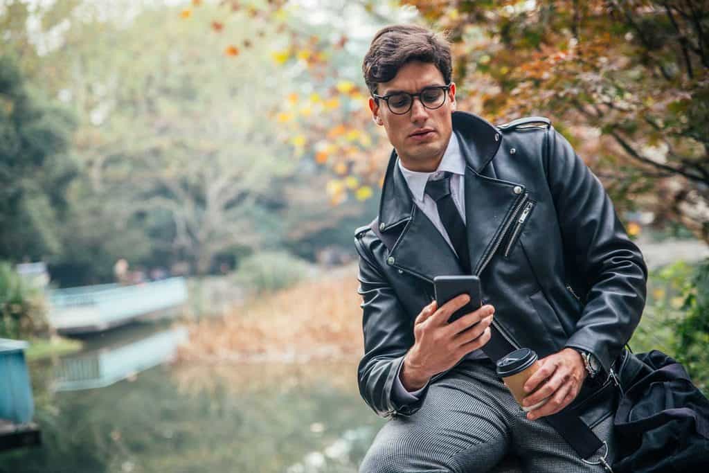 Young stylish businessman in leather jacket sitting in the park, text messaging and drinking coffee