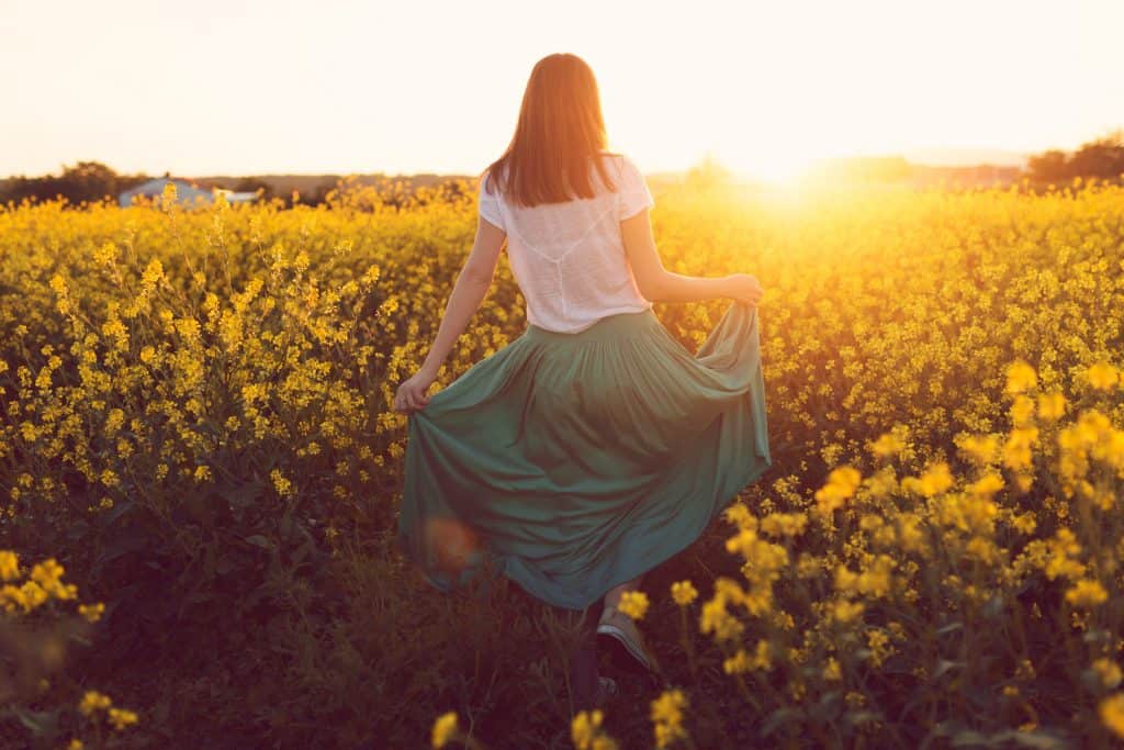 Rear view of a beautiful, russet young woman dancing in the middle of the flower meadow, surrounded by yellow flowers, spreading her skirt out