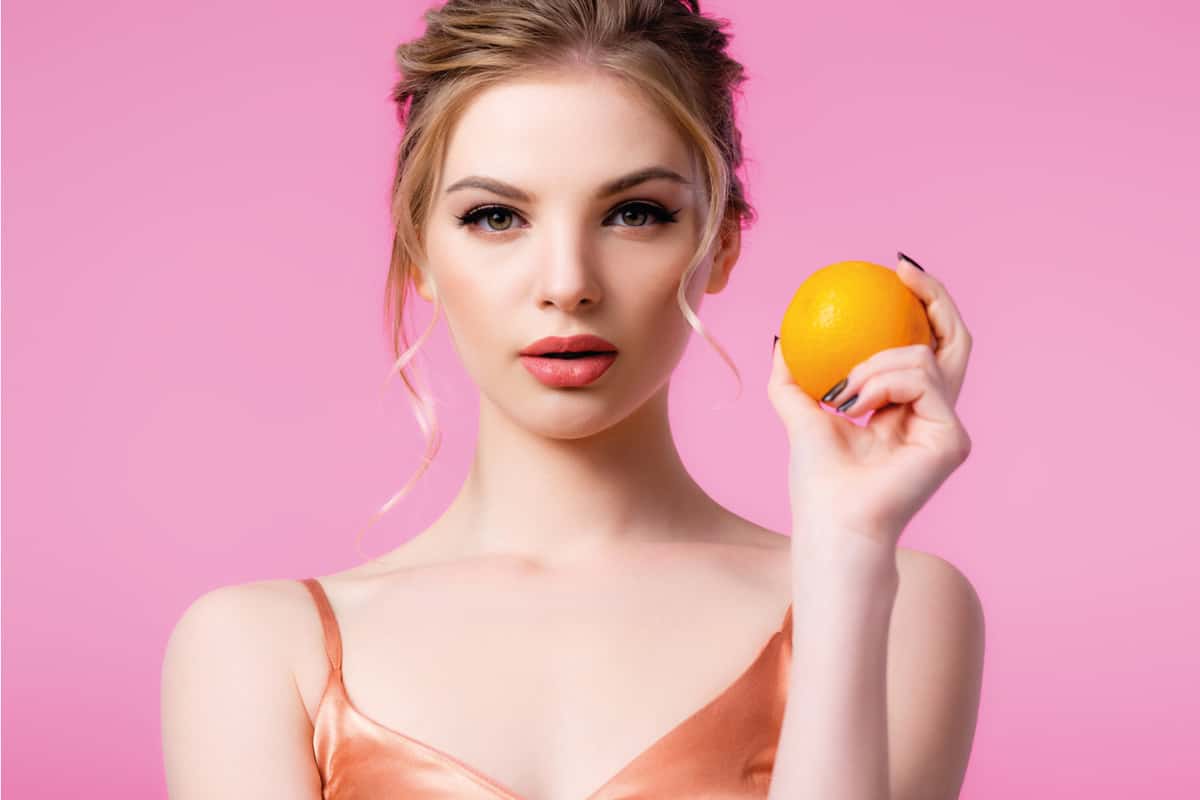 elegáns, gyönyörű szőke nő, aki érett narancsot tart, rózsaszínre izolálva, őszibarack színű rúzsot visel