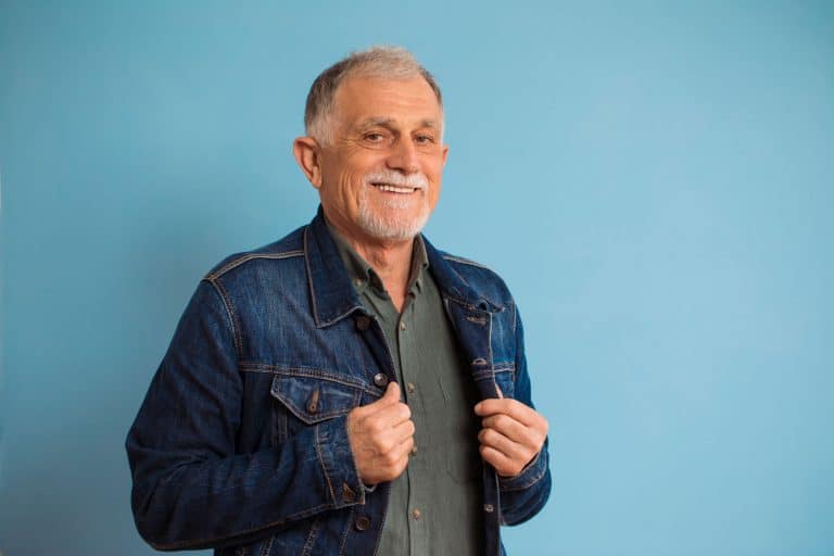 An elderly man tucking in his denim jacket for a photoshoot, How To Wear A Denim Jacket Over 50 [15 Awesome Ideas]