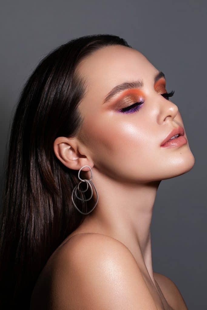 Close-up portrait of beautiful young brunette caucasian woman with colourful purple and orange eyeshadow. Shot in the studio on a plain grey background