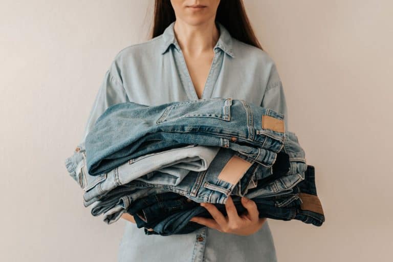 Unrecognizable woman holds stack of lot jeans pants on white background, How Often Should You Wash Pants? [A Breakdown By Type]