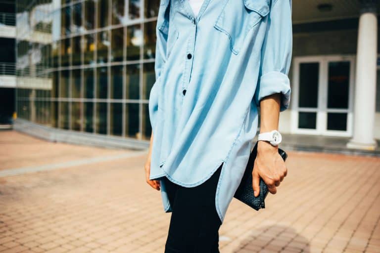 Unrecognizable young woman wearing oversize denim tunic shirt, black jeans and white wristwatches holding clutch bag standing outdoors in city on summer day, Can You Wear Tunics With Jeans? [With 6 Stylish Outfit Suggestions!]