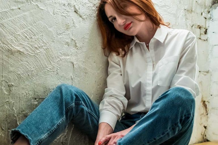 Woman with red lips in jeans and white shirt sits on wooden bench, What Color Lipstick Goes With Jeans? [A Look At Top Choices]