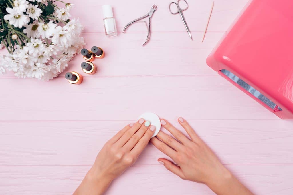 Top view floral composition female hands remove sticky layer from gel nail polish. Flat lay arrangement woman making shellac manicure herself near UV lamp, tools and bouquet of flowers on pink table.