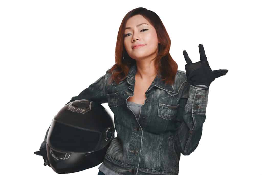 A lady rider wearing a denim jacket while wearing gloves and holding a a helmet on a white background