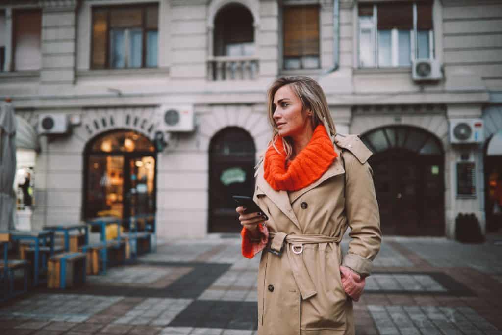 A tall blonde woman wearing a brown trench coat and holding her phone