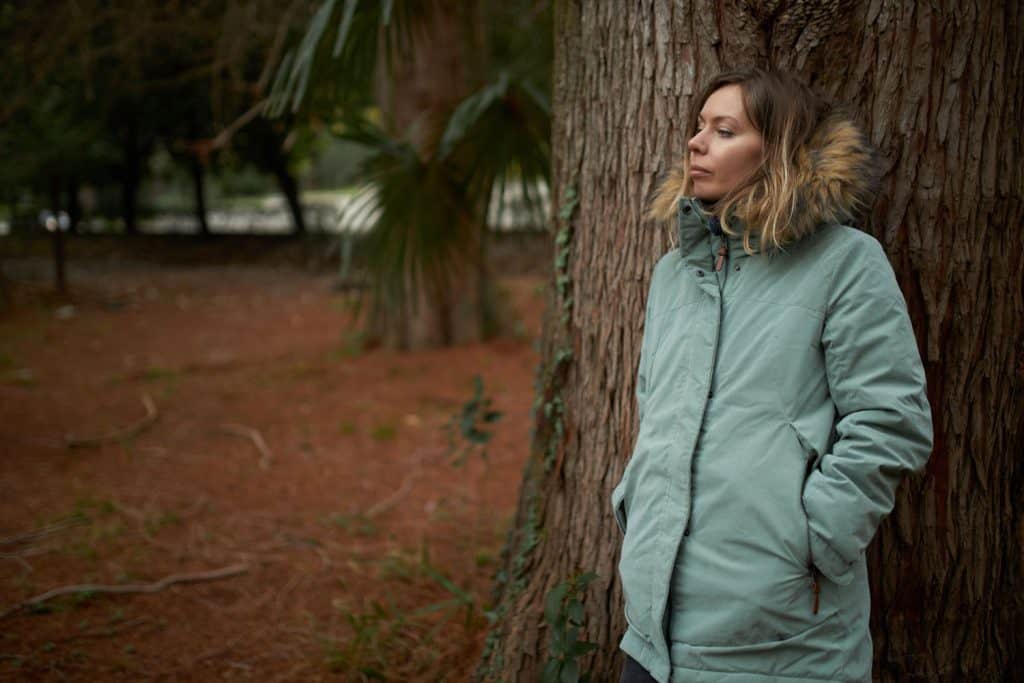 Good-looking young woman in green parka leaning against huge old tree isolated on blurred background of green exotic forest, having pensive face expression, stopping for break during hiking. Wearing parka
