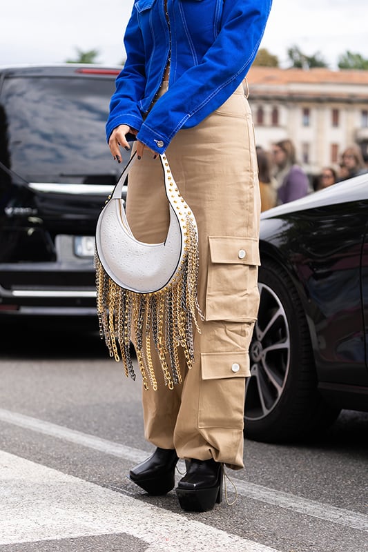 Woman in blue denim asymmetric cropped jacket, leather bag with nailed studded fringed chain pendant shoulder, beige cargo flared pants, platform high heels shoes