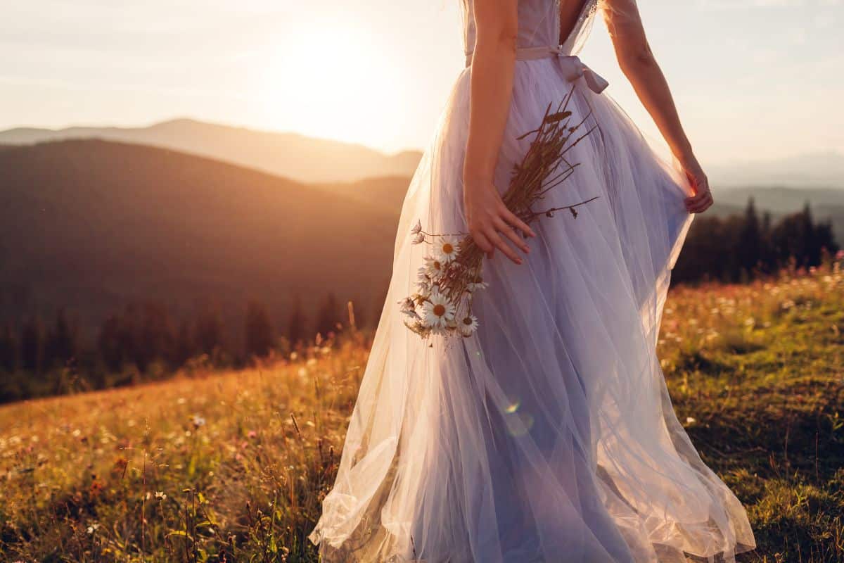Bride wearing blue wedding dress holding bouquet in mountains at sunset. Woman walking on meadow in flowers. Fashion model posing in nature.