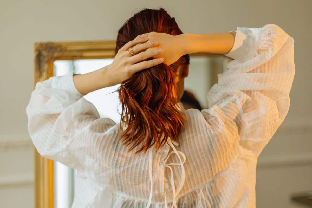A beautiful red-haired girl in the bathroom looks in the mirror and ties her ponytail. Pretty woman stroking her hair while looking in the mirror. The concept of hairstyles and hair care. Back view