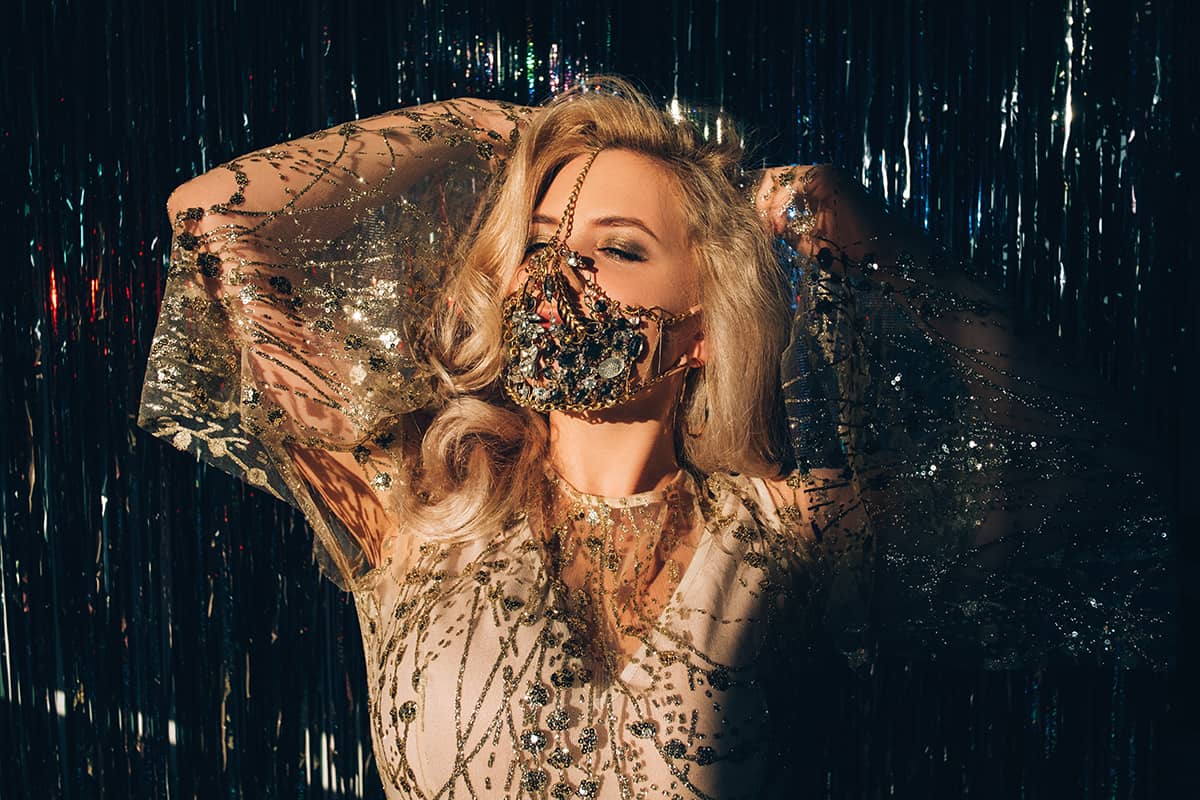 Mysterious blonde woman in luxury chain face mask matching elegant dress posing on dark shiny tinsel background