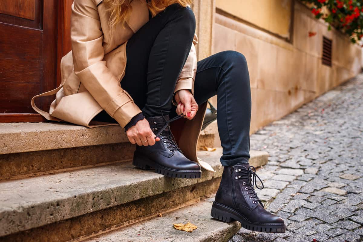 Woman wearing trench coat sitting on staircase and tying shoelace on her ankle boot.