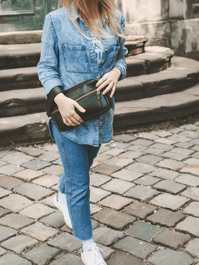 Fashionable blonde in denim shirt, jeans, white sneakers and black leather handbag walking around the city