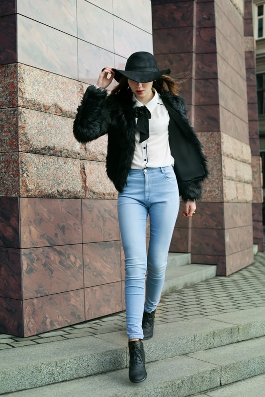 Full body portrait of a young beautiful lady walking at street of the old city. Model wearing stylish black fur coat and wide-brimmed hat.