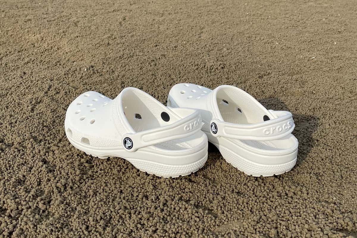 A pair of white Crocs footwear on a sandy beach. Branded foam clog.
