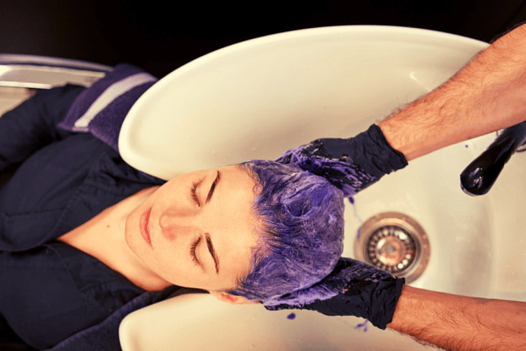 Image of a woman over a shampooing bowl sink and two hands rubbing toner into her hair.