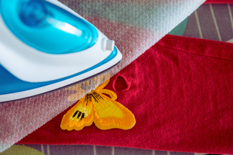 Image is of an iron on an ironing board. Between the iron and ironing board is an iron-on patch and a piece of fabric. The patch is in the shape of a butterfly.