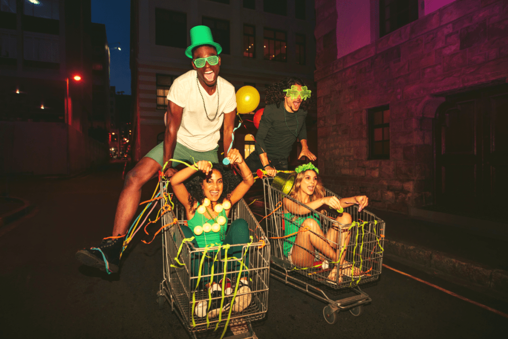 Photo is of a group of friends on St. Patrick's Day. Two individuals are riding in shopping carts down a street while the other two are pushing. They appear to be having a great time.