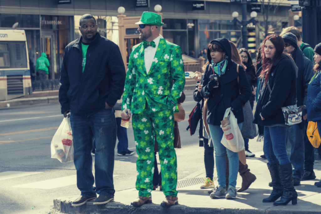 Photo of a crowd waiting at a light to cross the street. One man is dressed in a St. Patrick's Day suit. Onlookers are looking at his suit in a funny way.