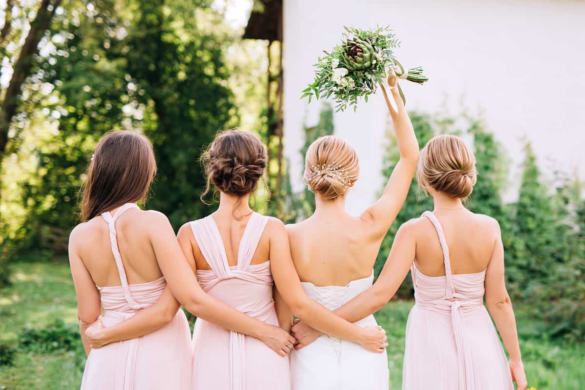 bride standing with her back in a white wedding dress and raised hand with three bridesmaids
