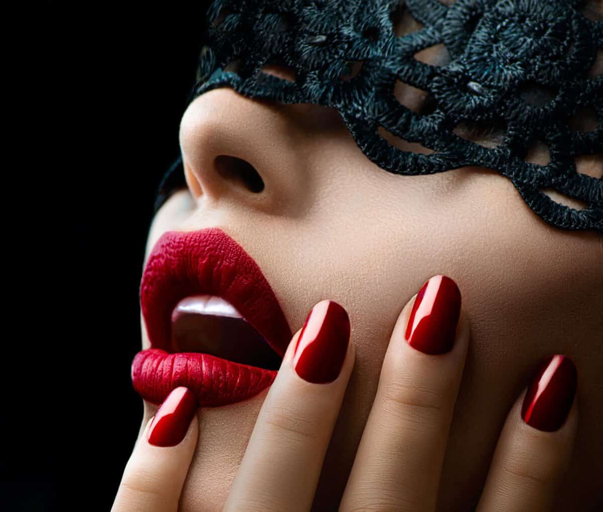 Beautiful Woman with Black Lace mask over her Eyes. Red Sexy Lips and Nails closeup. 
